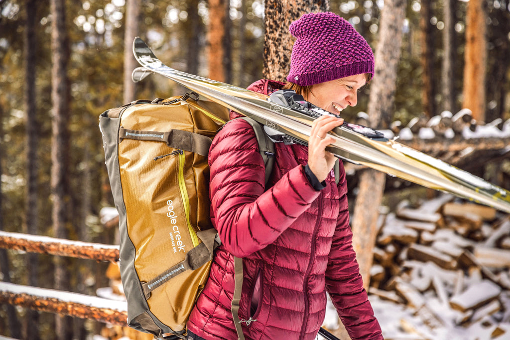 A woman carries skis on her shoulder and a duffel bag on her back